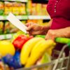 Persona revisando una lista de la compra junto a un carrito con frutas y alimentos frescos en un supermercado.