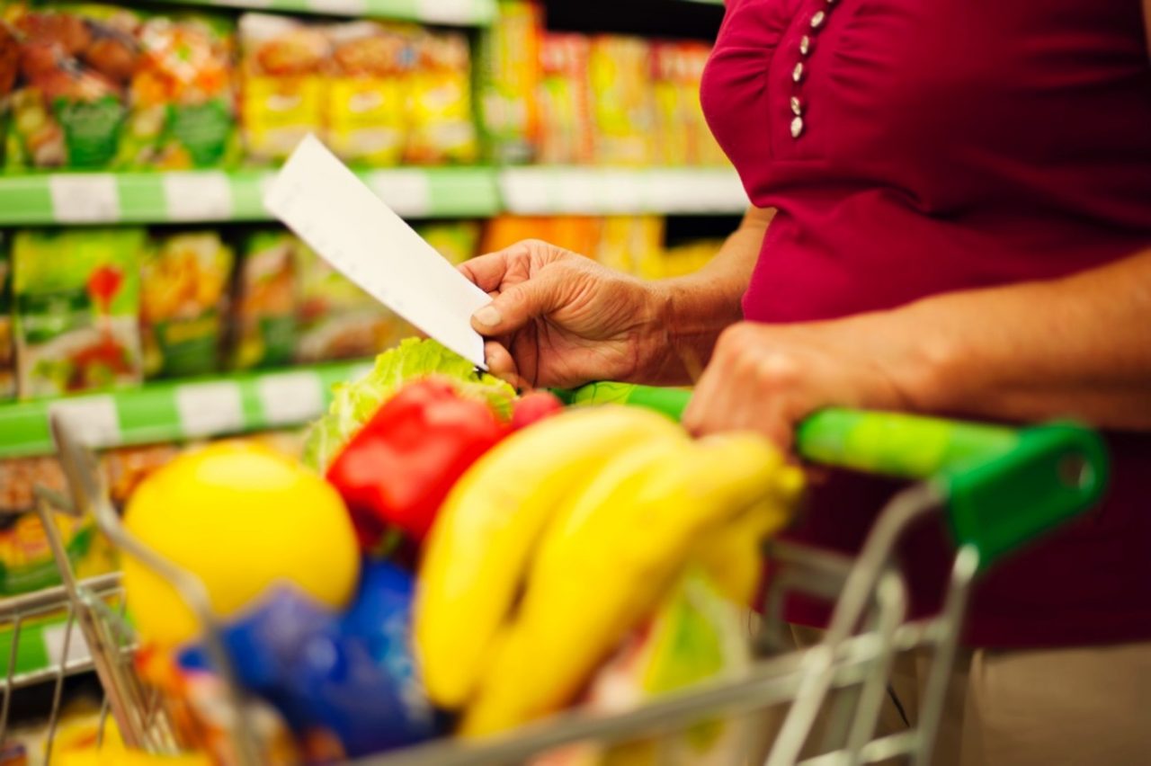 Persona revisando una lista de la compra junto a un carrito con frutas y alimentos frescos en un supermercado.