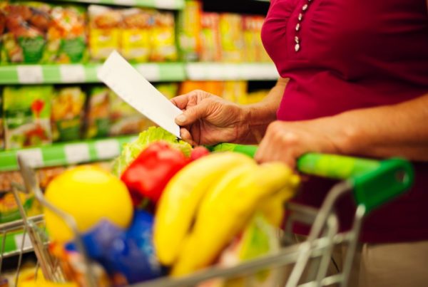 Persona revisando una lista de la compra junto a un carrito con frutas y alimentos frescos en un supermercado.