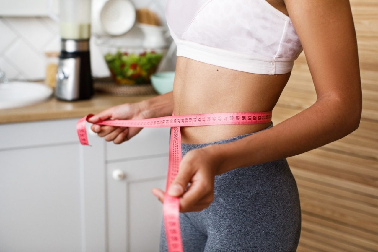 Mujer midiendo su cintura con una cinta métrica en la cocina, relacionada con estrategias nutricionales para perder grasa de forma saludable.