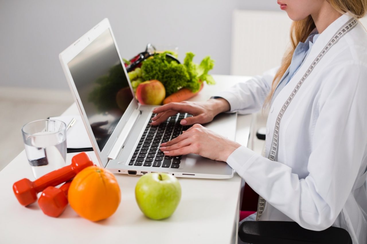 Nutricionista trabajando con un ordenador portátil junto a frutas y verduras, representando la consulta de nutrición online y presencial.